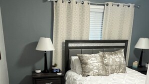 Une chambre à coucher avec un lit, deux tables de chevet avec des lampes, une horloge et une fenêtre avec des rideaux.
