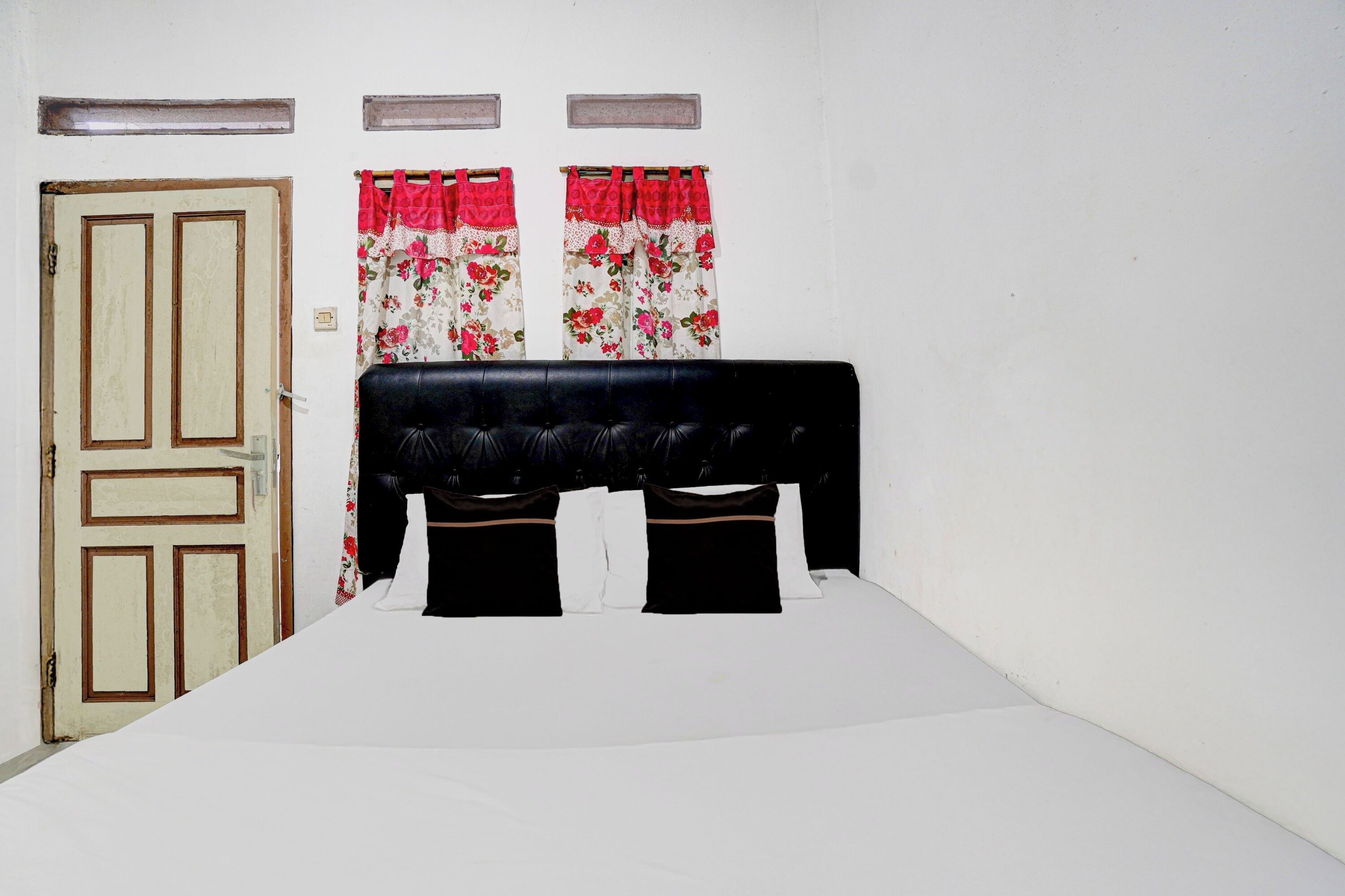 A bed with a black headboard, two windows with floral curtains, and a wooden door.