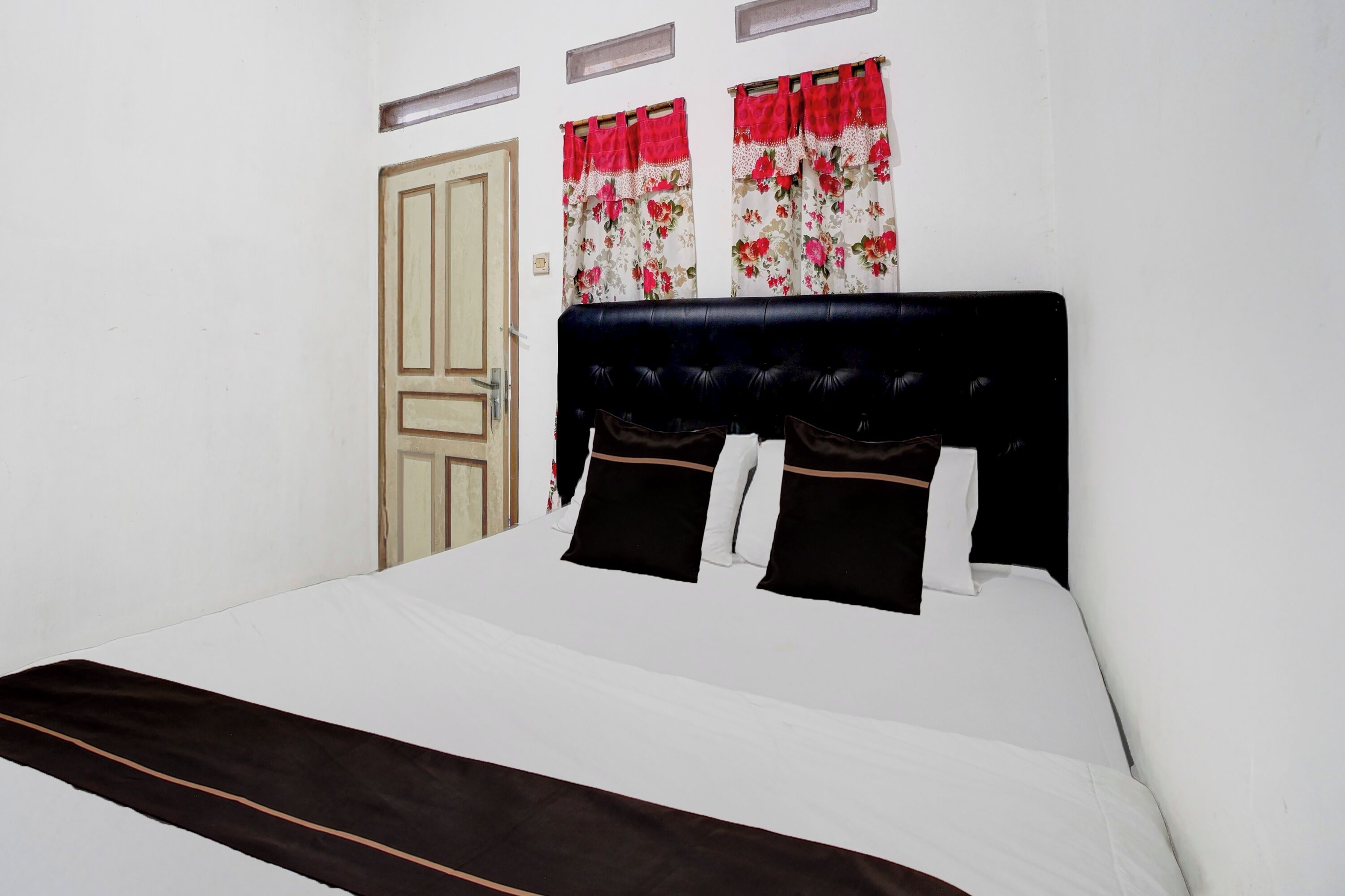 Een netjes opgemaakt bed met een zwart hoofdbord en witte beddengoed, een houten deur en bloemen gordijnen.