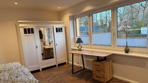 Une chambre à coucher comprenant un lit, un bureau, une armoire et une fenêtre donnant sur des arbres et un immeuble.