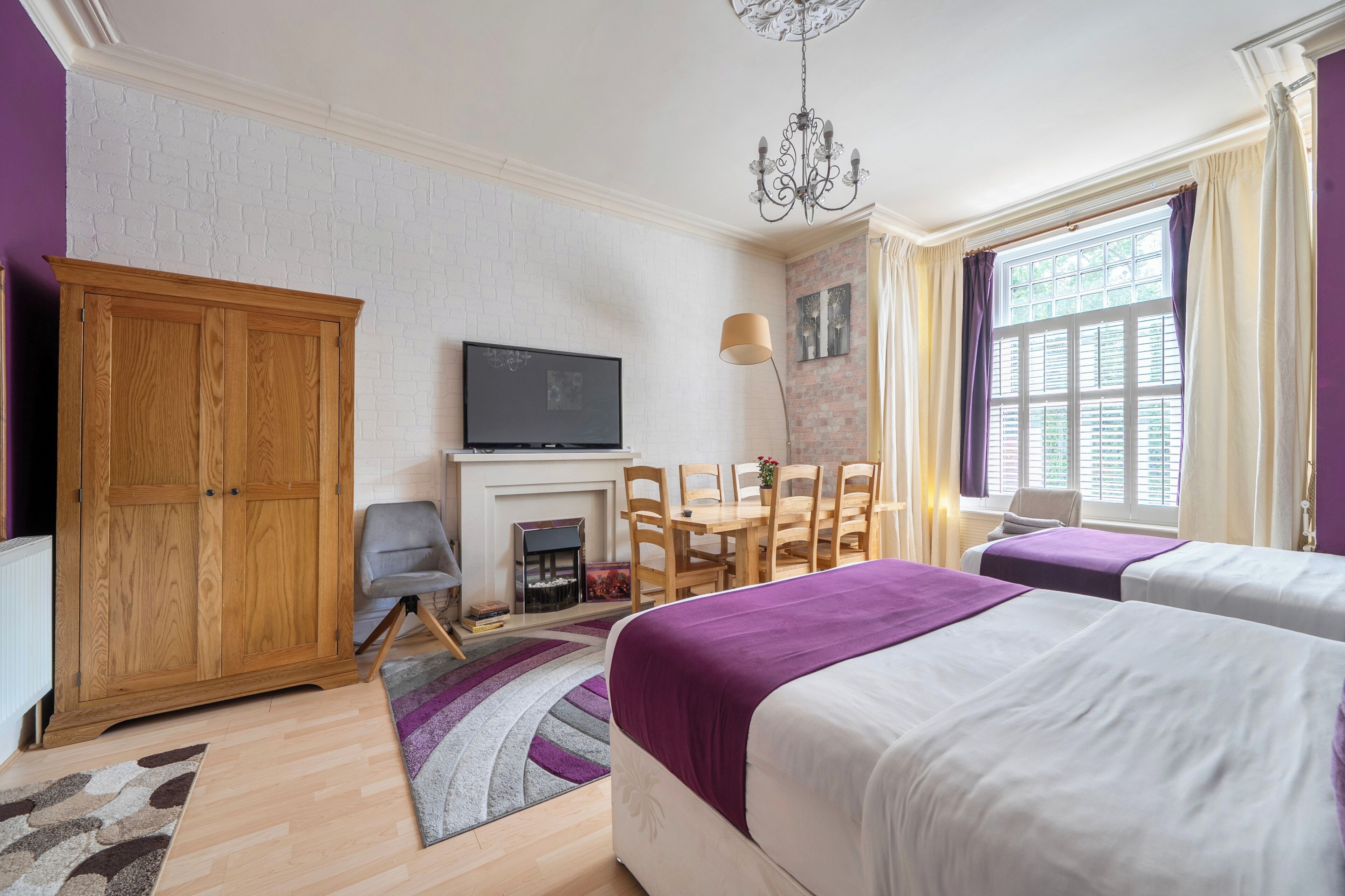A bedroom with two beds, a TV, a fireplace, a dining table with chairs, and a large window with curtains.