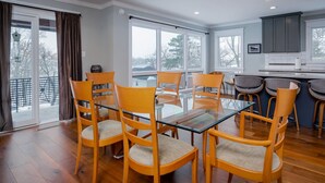 A dining area with a glass table and wooden chairs, a kitchen with grey cabinets, and large windows with a view of trees.