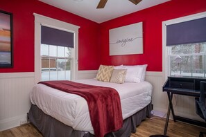 Slaapkamer met een rode wand, wit beddengoed, een bed met een rode plaid en een bureau met een lamp.
