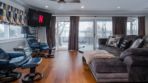A modern living room with a large flat-screen TV, leather chairs, a grey sofa, and a balcony with a view.