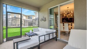 A modern living room with a white sofa, a small table, and a large sliding glass door leading to a kitchen and outdoor area. - Pool & BBQ - Disney-Themed Cozy 6BD - Near Disney (Kissimmee)