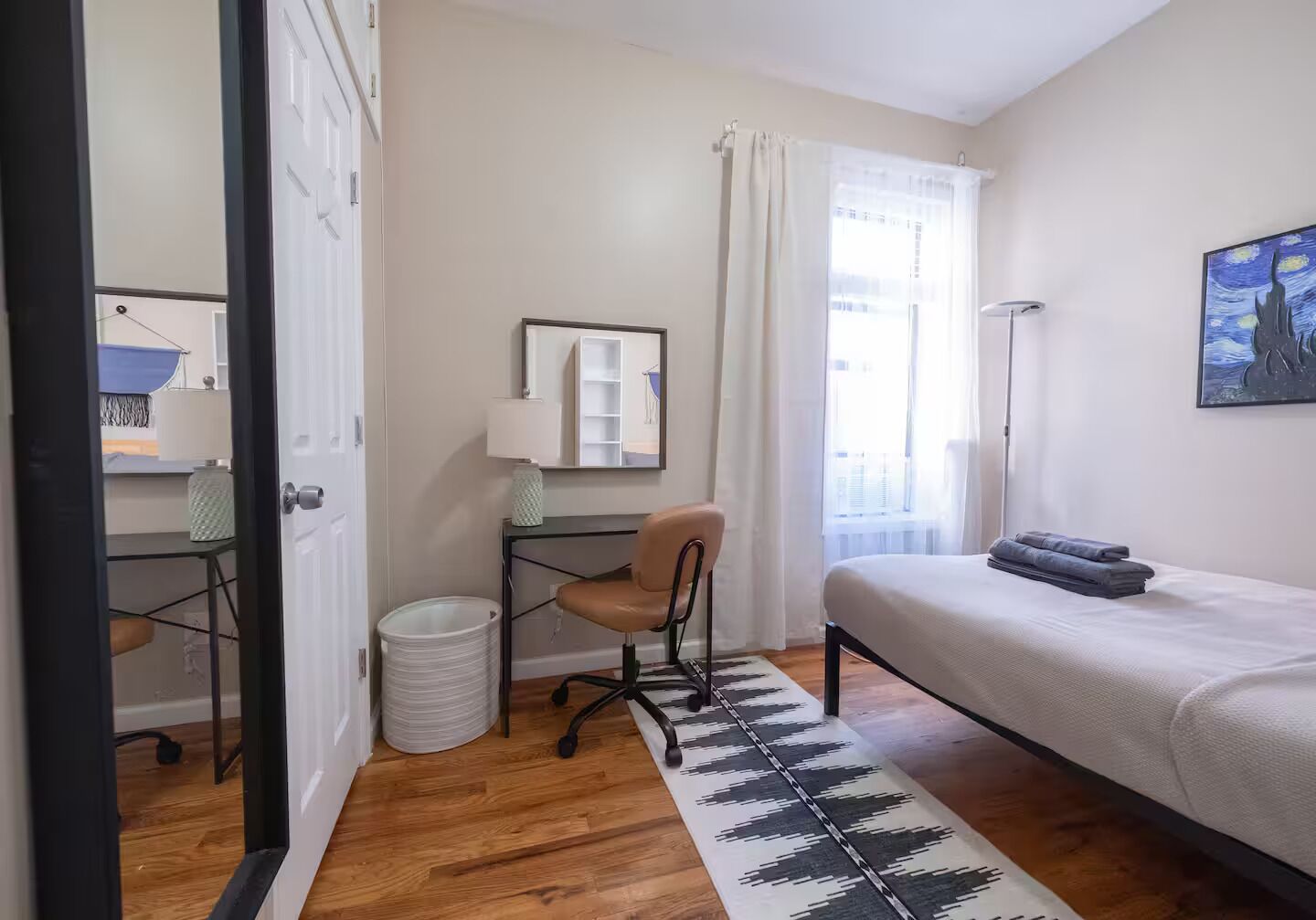 A bedroom with a bed, a desk, a chair, a mirror, a lamp, a window with curtains, and a painting on the wall.