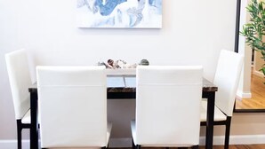 Uma sala de jantar com uma mesa de madeira e cadeiras brancas, um quadro abstrato grande na parede e um vaso com planta no canto.