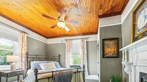 Een slaapkamer met een houten plafond, een bed met metalen frame, een nachtkastje, een lamp, een raam met gordijnen en een open haard.