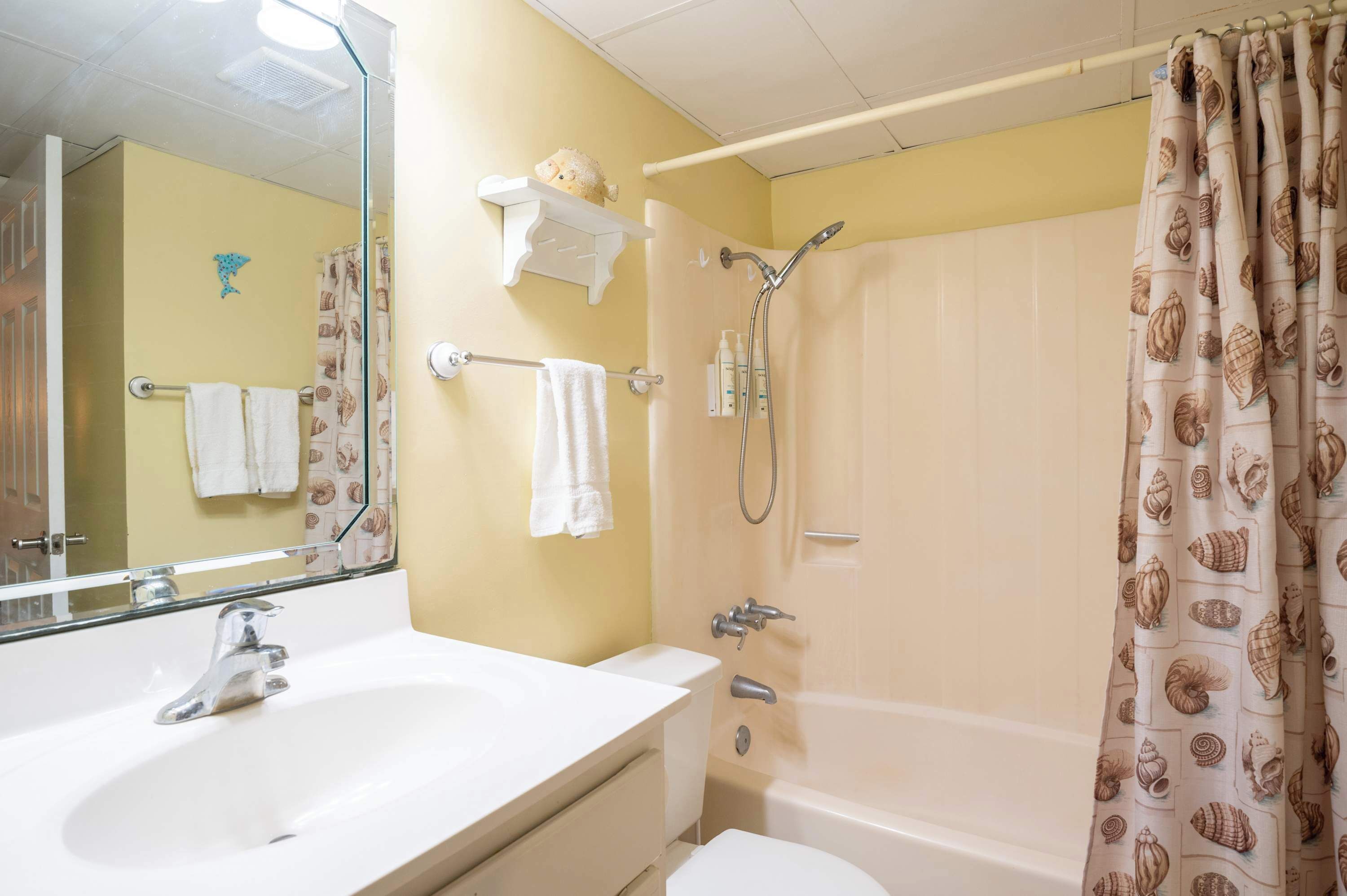 A bathroom with a white sink, toilet, and bathtub. There is a towel rack with two white towels and a shower curtain with a shell pattern.