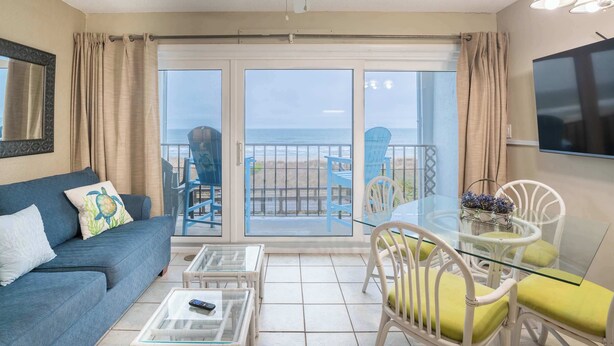 Ett vardagsrum med en soffa, ett soffbord och ett matbord med stolar. Det finns en balkong med utsikt över havet och strandstolar.
