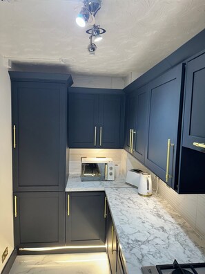 Une cuisine moderne avec des meubles de couleur bleu foncé, un plan de travail en marbre blanc, un four à micro-ondes encastré, un grille-pain et une cuisinière à gaz.