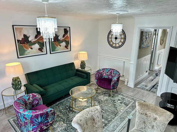 Un salon avec un canapé vert, deux fauteuils à motifs, une table basse et un lustre.