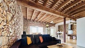 Un salon avec un mur en pierre, des poutres apparentes en bois au plafond, un canapé avec des coussins, une table basse en bois et, au fond, un coin cuisine.