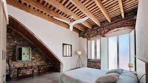 Une chambre avec un plafond en pente, un lit recouvert d’une courtepointe, une lampe de chevet et un mur en pierre avec une fenêtre.
