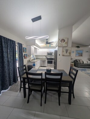 Un coin repas avec une table et des chaises, un ventilateur de plafond, et une cuisine en arrière-plan.