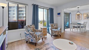 A modern living room with a large window, a dining area with white chairs, and a small table. - Sun & Fun | Beachfront Resort~Pool~Lazy River~Gym (Myrtle Beach)