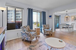 A modern living room with a large window, a dining area with white chairs, and a small table. - Sun & Fun | Beachfront Resort~Pool~Lazy River~Gym (Myrtle Beach)
