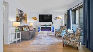 A living room with a sofa, chairs, a TV, a fireplace, and a large window with curtains. - Sun & Fun | Beachfront Resort~Pool~Lazy River~Gym (Myrtle Beach)