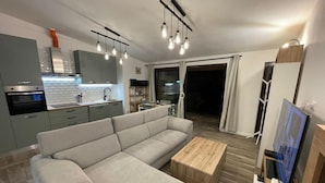 Un soggiorno moderno con un divano angolare grigio, un tavolino in legno e una TV su un mobiletto in legno. È presente un angolo cottura con mobili grigi e forno a incasso. La stanza ha un'ampia finestra con tende e un lampadario.