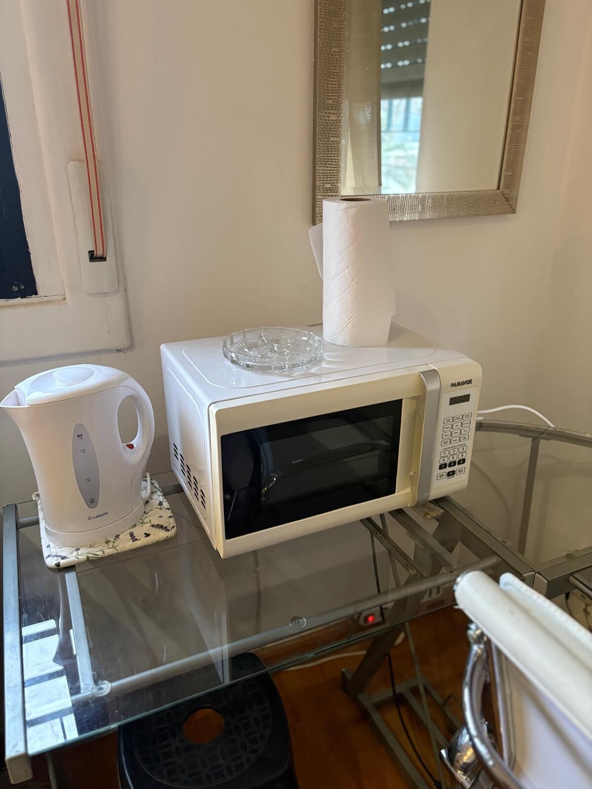 A microwave on a glass table with a kettle and a roll of paper towels.