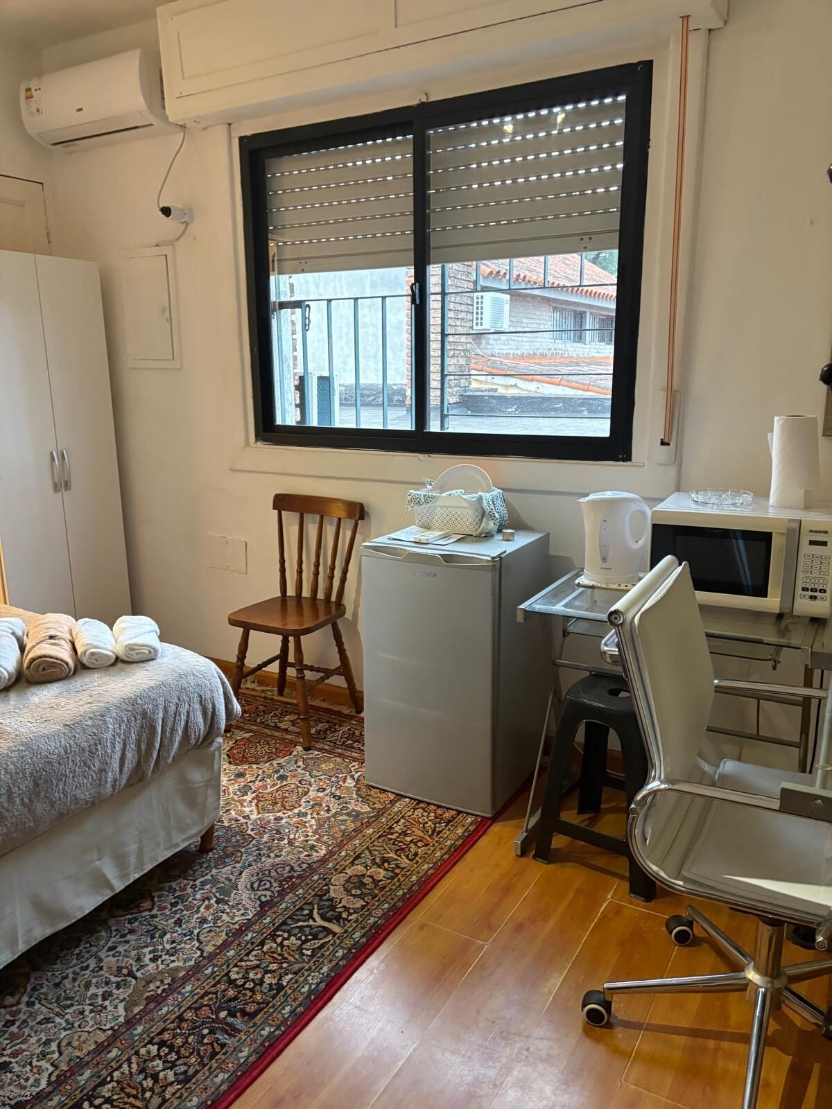 Une chambre d’hôtel compacte comprenant un lit, un petit bureau avec un micro-ondes, une chaise et une fenêtre donnant sur des immeubles.