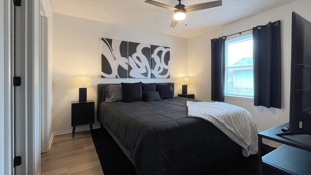 A bedroom with a bed, two bedside tables, a ceiling fan, a window with curtains, and a wall-mounted TV.
