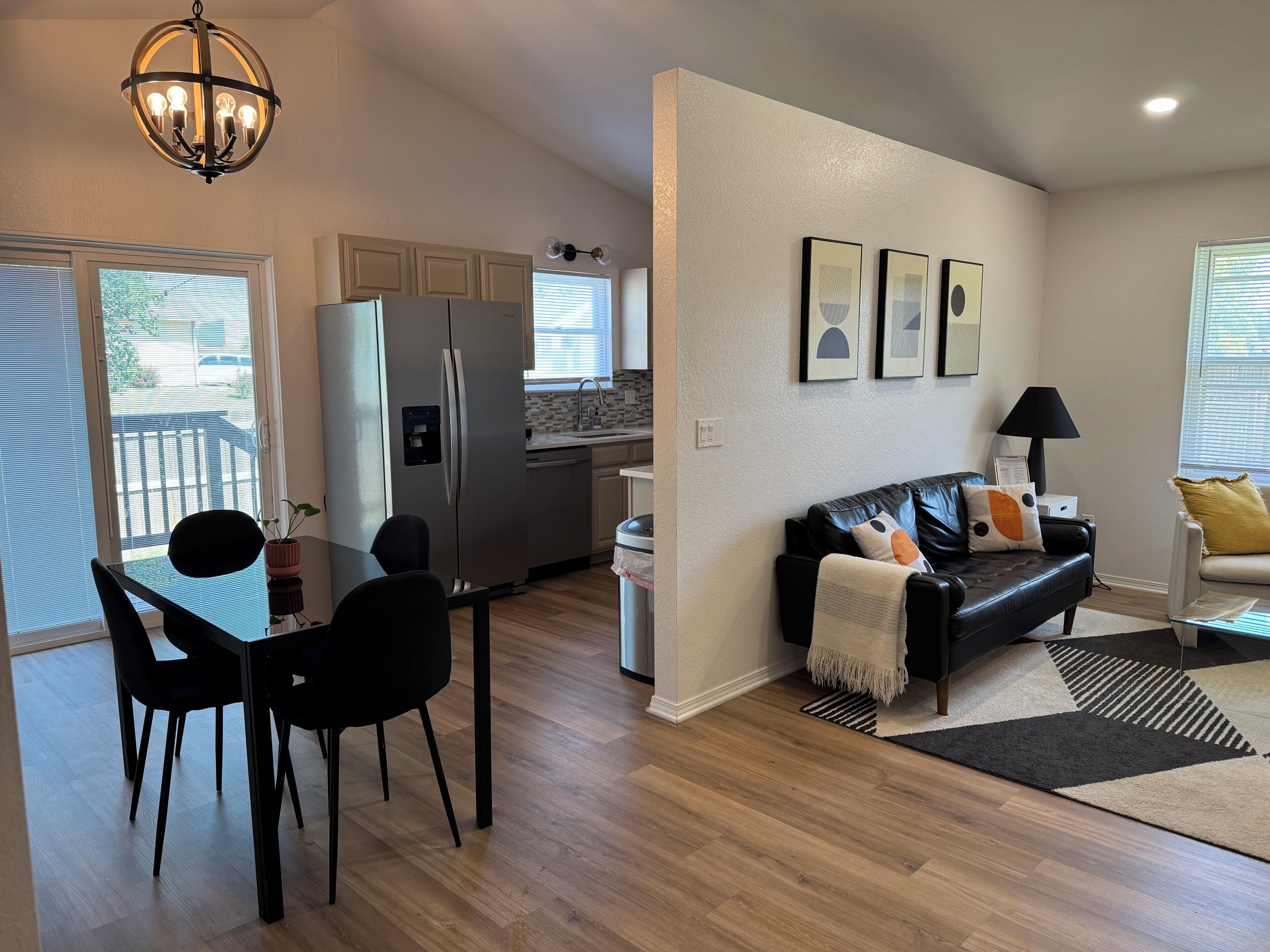 A modern kitchen with a refrigerator, a dining area with a table and chairs, a black leather sofa, and a chandelier.