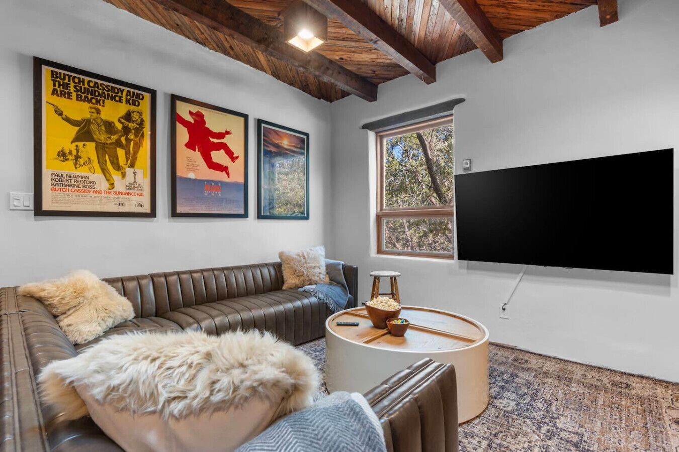 Ein Wohnzimmer mit braunem Ledersofa, einer weißen Kunstfelldecke, einem Couchtisch mit einer Schüssel Popcorn, einem großen Flachbildfernseher und gerahmten Filmplakaten an der Wand.