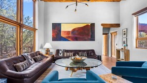 A spacious living room with a large window, a brown leather sofa, a blue armchair, a glass coffee table, and a painting of a mountain landscape. - Authentic Adobe Retreat - Family & Group Getaway (Santa Fe)