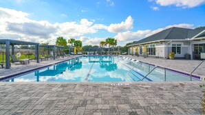 A swimming pool with lounge chairs and umbrellas, surrounded by a paved deck and a building with a grey roof. - Beautiful 3BR Ocala Home | Perfect for Families (Ocala)