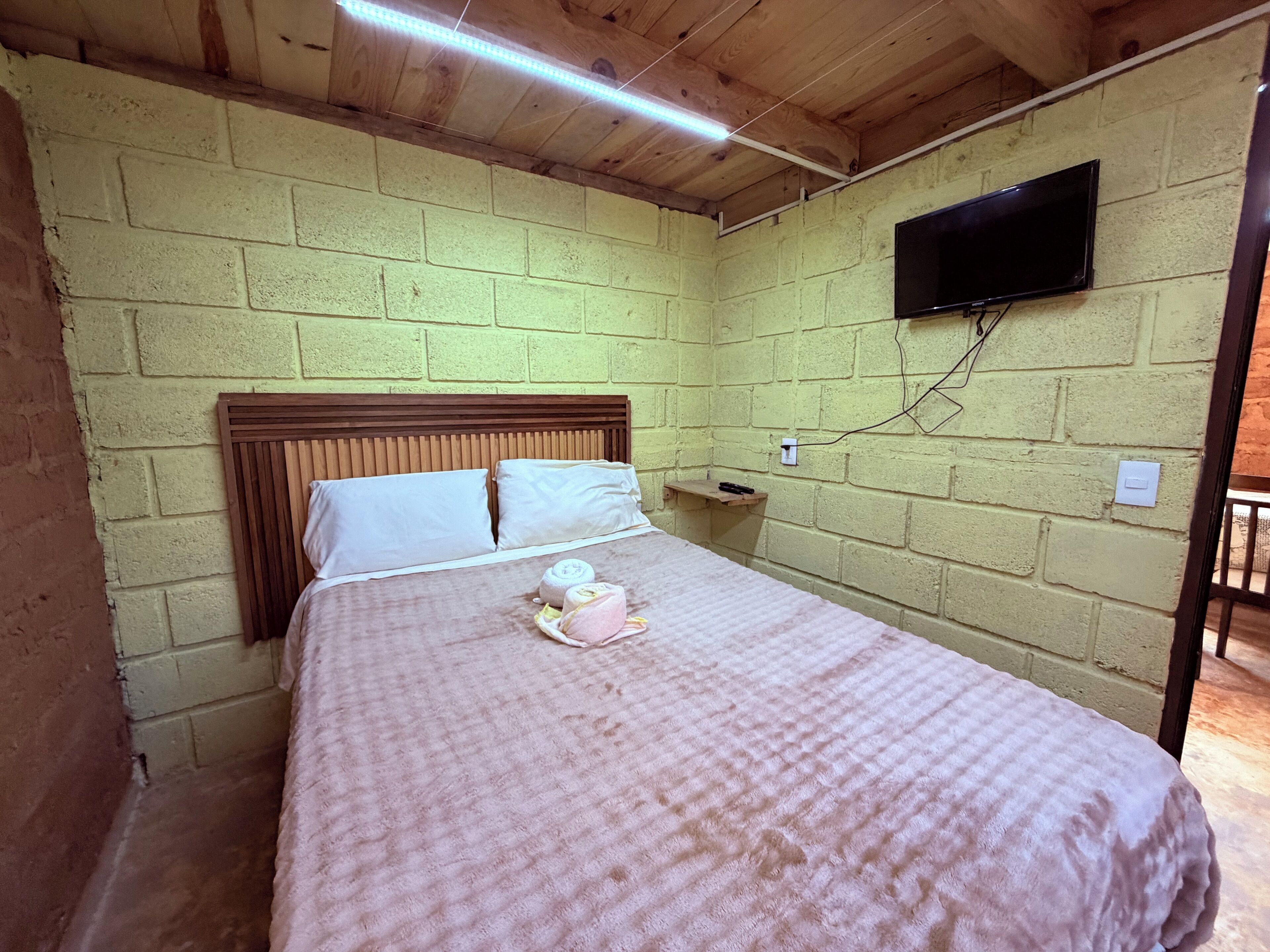 A small, single-bed room with a wooden headboard, a flat-screen TV mounted on the wall, and a small shelf.