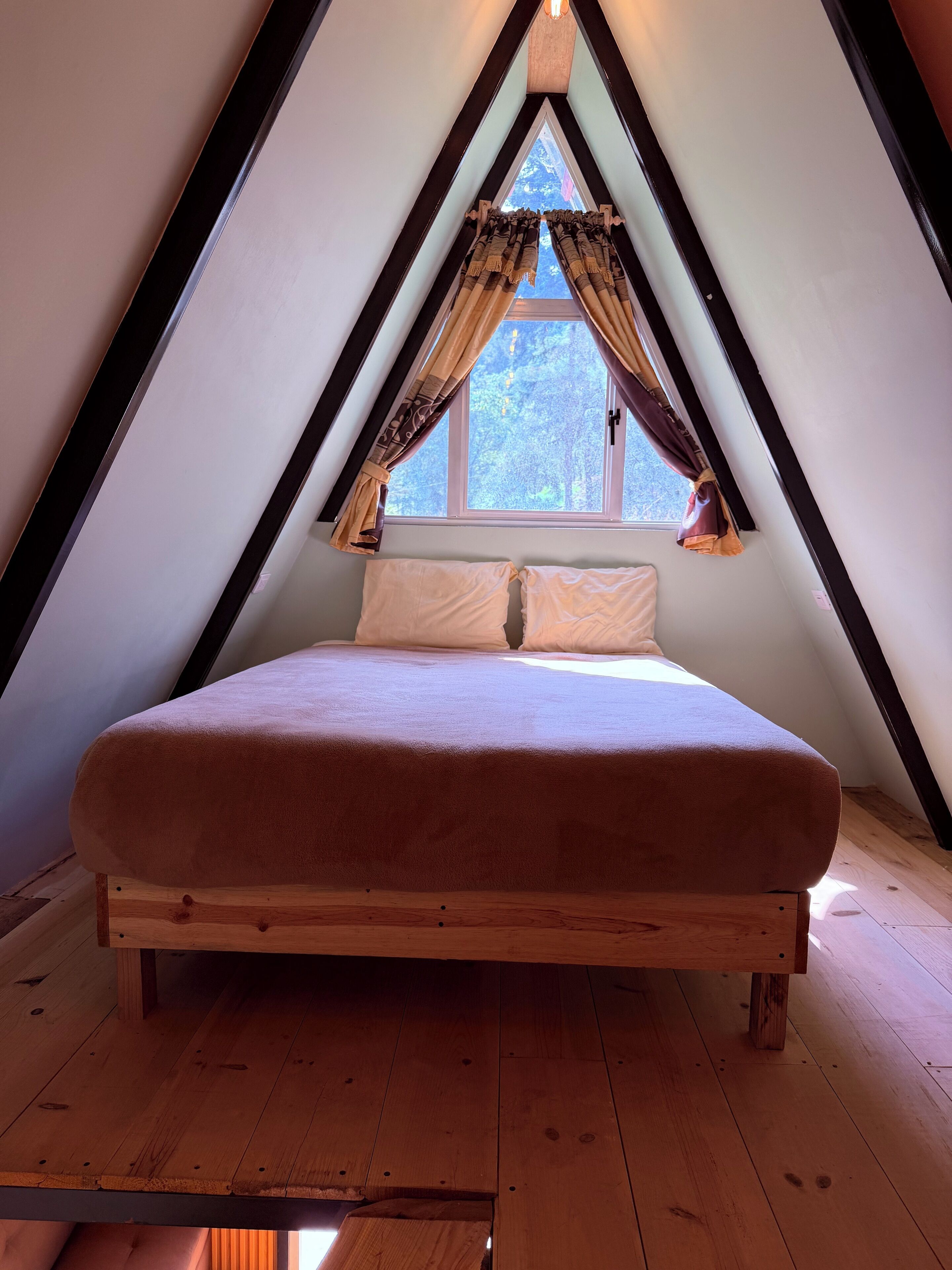 Ein Schlafzimmer mit Bett in einem giebelartigen Dachraum, Holzboden und einem fenster mit Vorhängen.