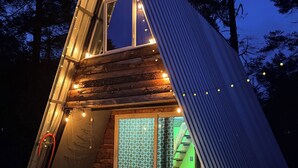 Una estructura metálica triangular con interior de madera, luces colgantes y una puerta de vidrio iluminada en verde.