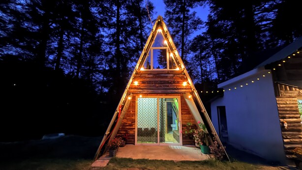 Una cabaña de madera triangular con puerta y ventanas de vidrio, rodeada de árboles.