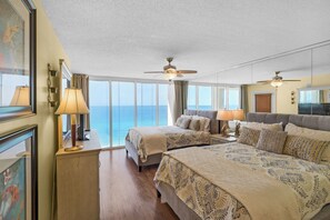 Ein Hotelzimmer mit zwei Betten, einem großen Fenster mit Meerblick, Deckenventilatoren und einem Holzboden.