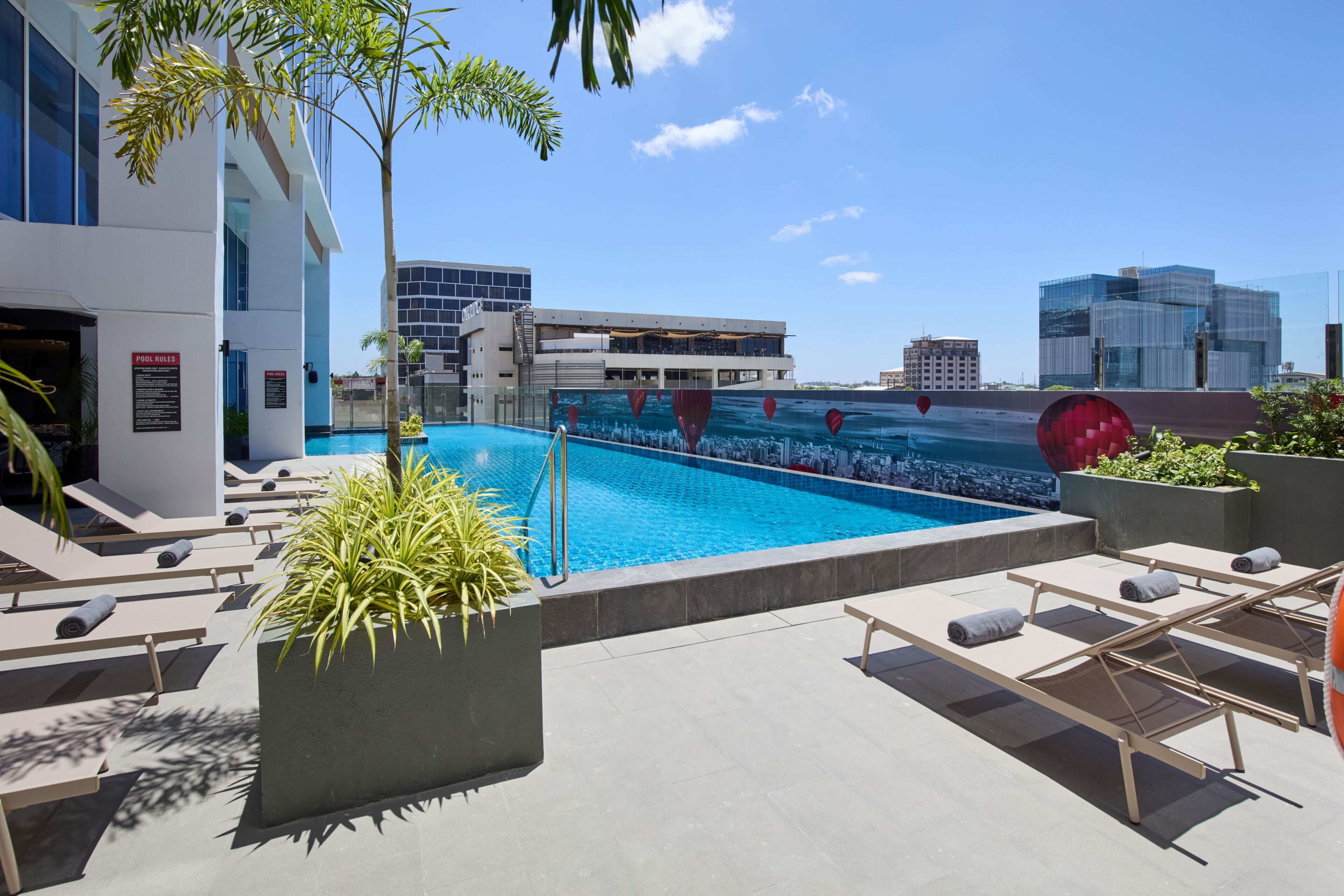 Una piscina en la azotea con sillones, un edificio moderno al fondo y un cielo despejado.