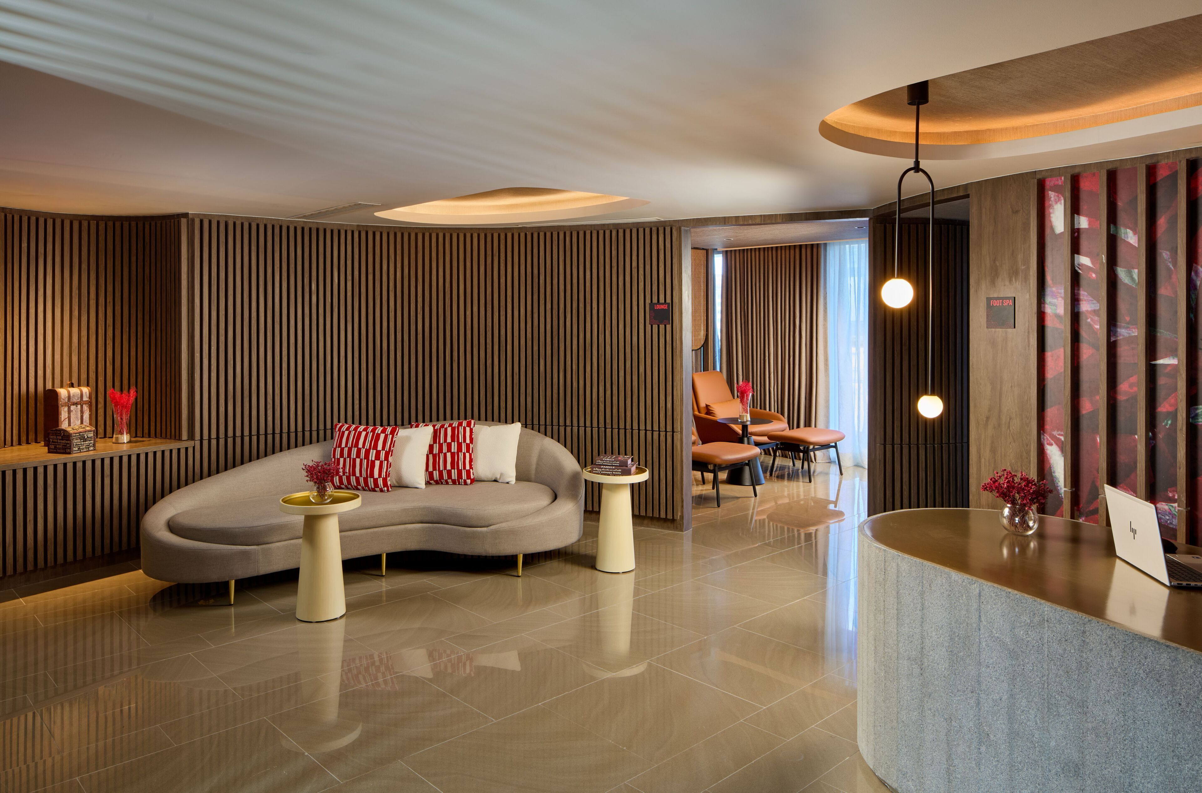 A modern lobby with a curved reception desk, a sofa with red and white cushions, and a small side table with a vase.