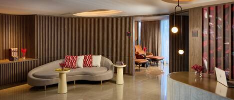 A modern lobby with a curved reception desk, a sofa with red and white cushions, and a small side table with a vase.