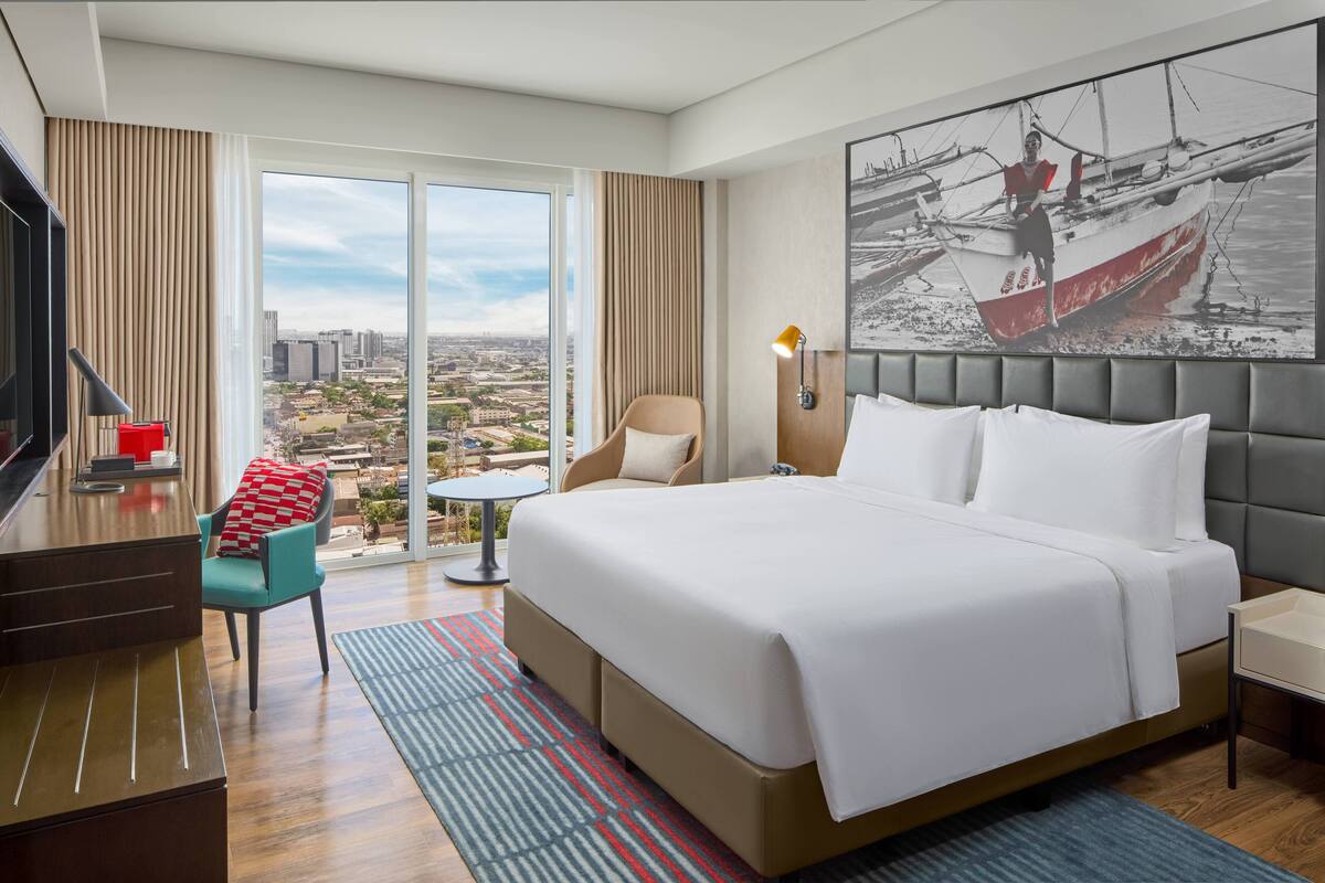 A hotel room with a large bed, a desk, a chair, a small table, and a view of the city.