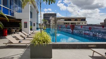 A rooftop pool with sun loungers, a bar area, and modern buildings in the background.