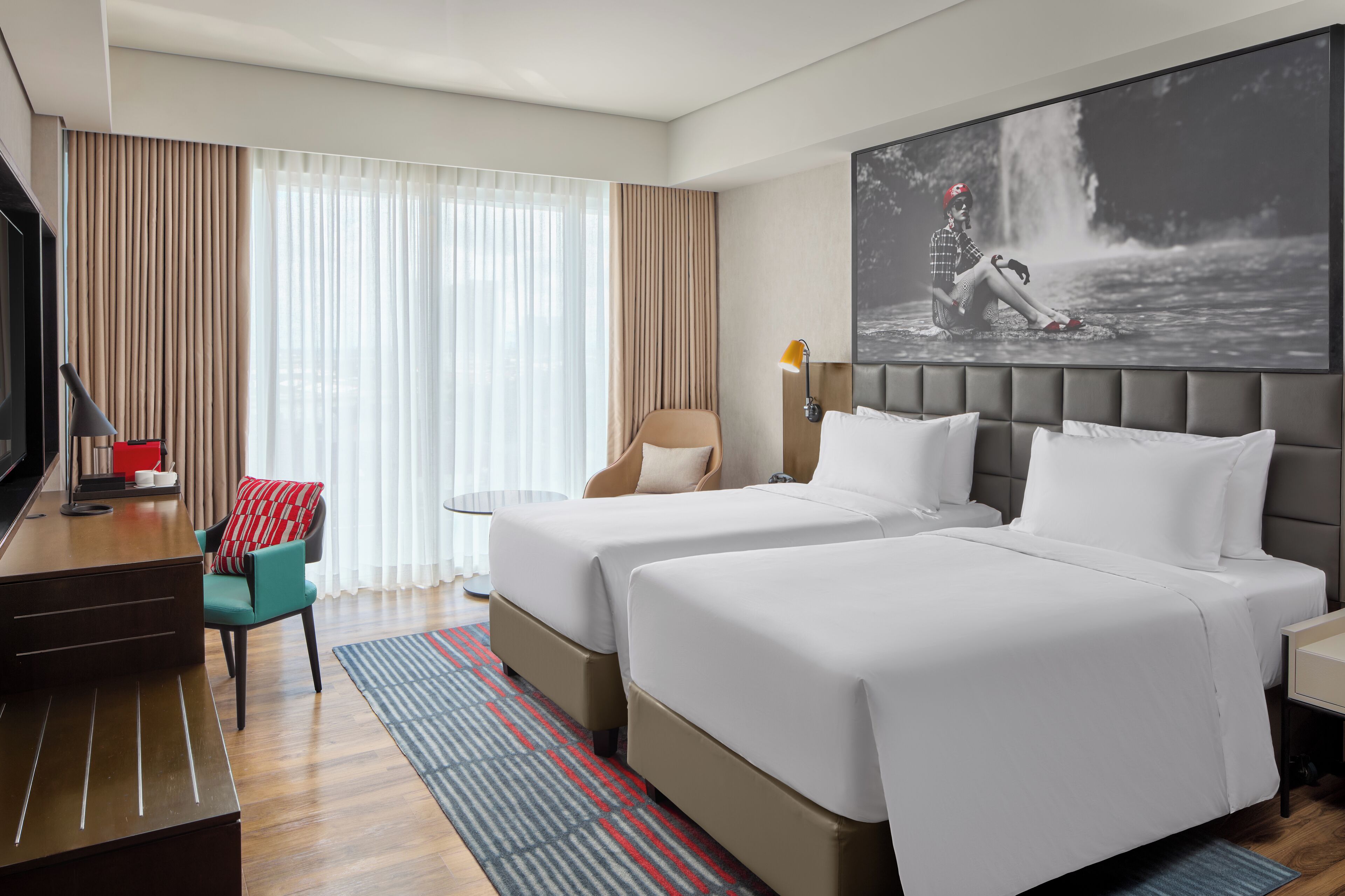 A hotel room with two beds, a desk, a chair, and a large window with curtains.