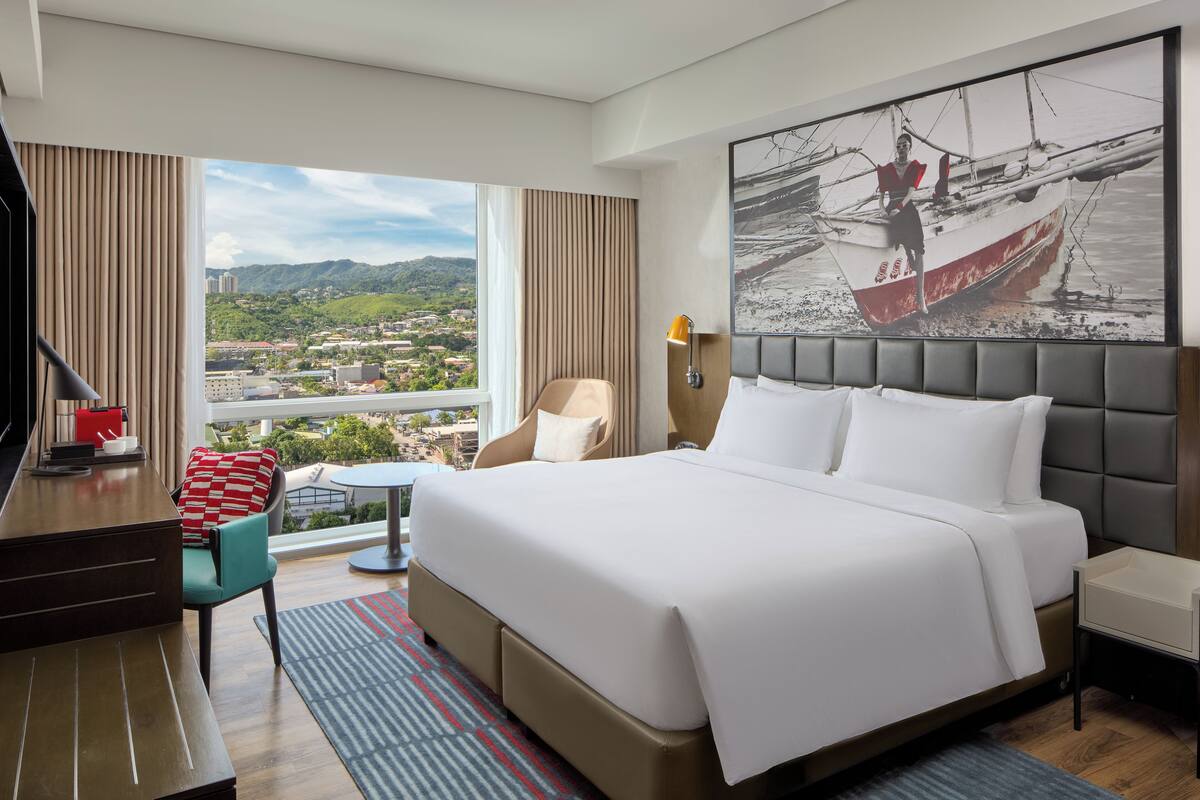A hotel room with a large bed, a desk, a chair, a small table, and a view of a cityscape through large windows.