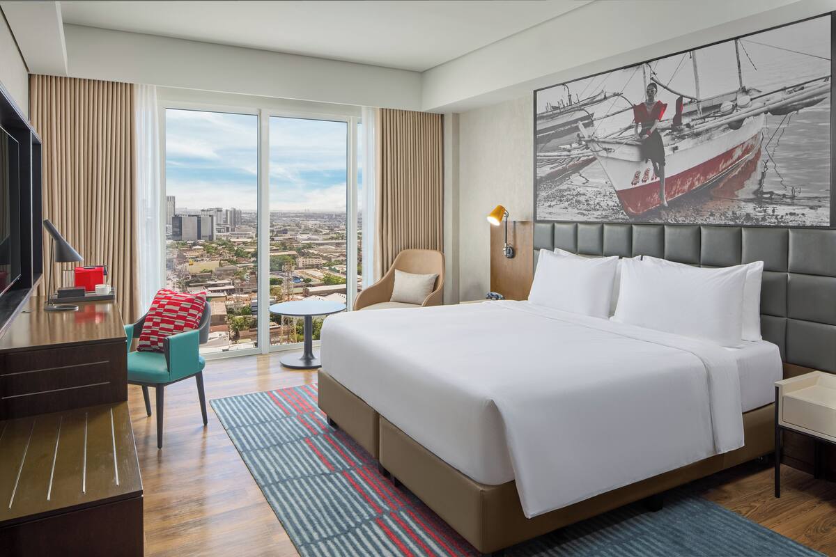 A hotel room with a large bed, a desk, a chair, a small table, and a large window with a city view.