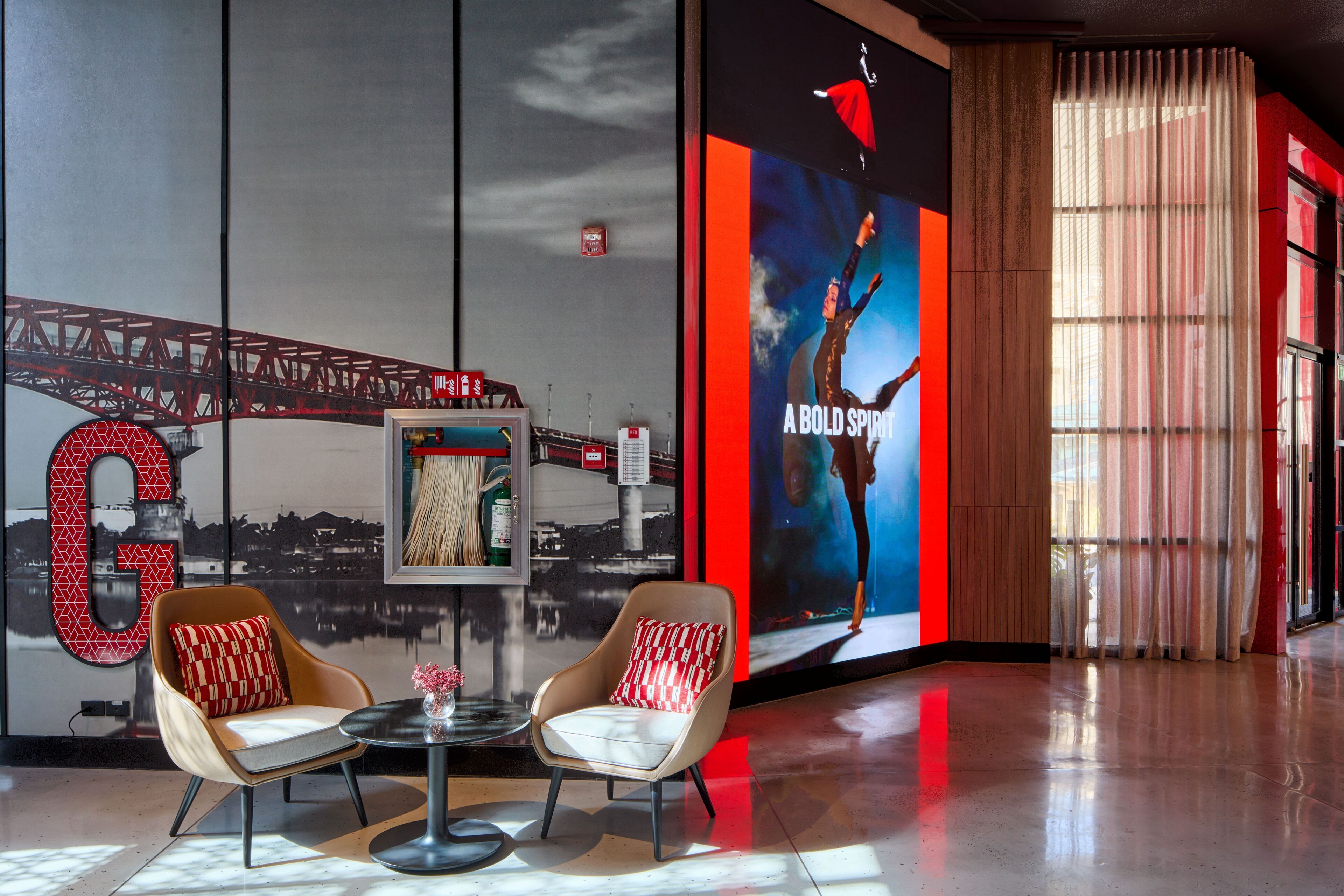A modern lobby with two armchairs, a round table, a large digital screen displaying a cityscape, and a red and white poster.