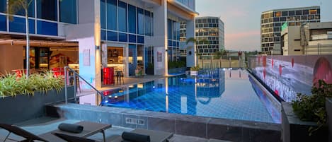 Una piscina en la azotea con sillones de descanso, un restaurante y edificios modernos al fondo.