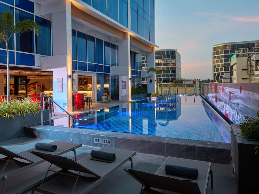 A rooftop pool with lounge chairs, a restaurant, and modern buildings in the background.
