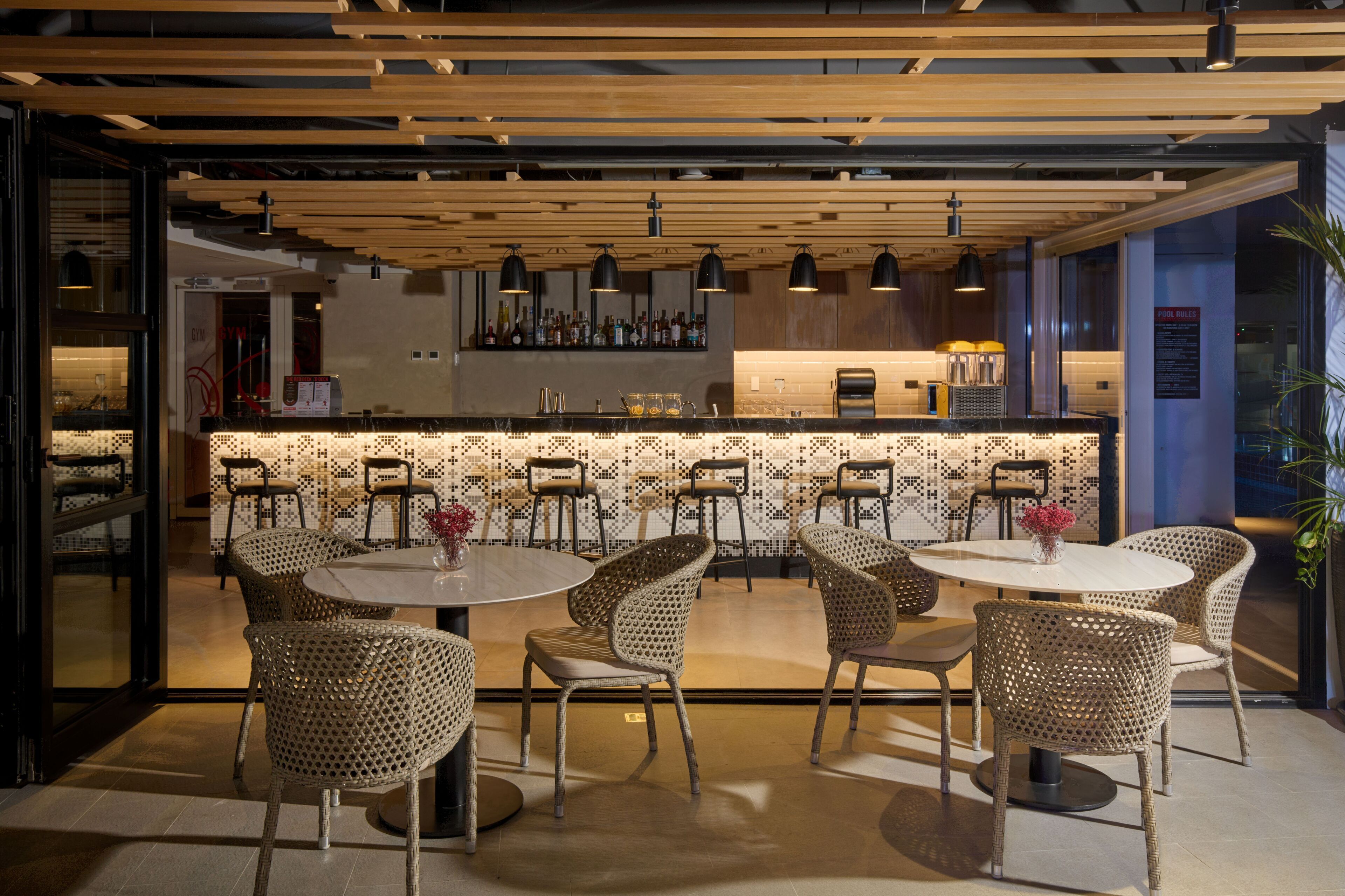 A restaurant with round tables and wicker chairs, a bar area with bottles, and a modern ceiling design.