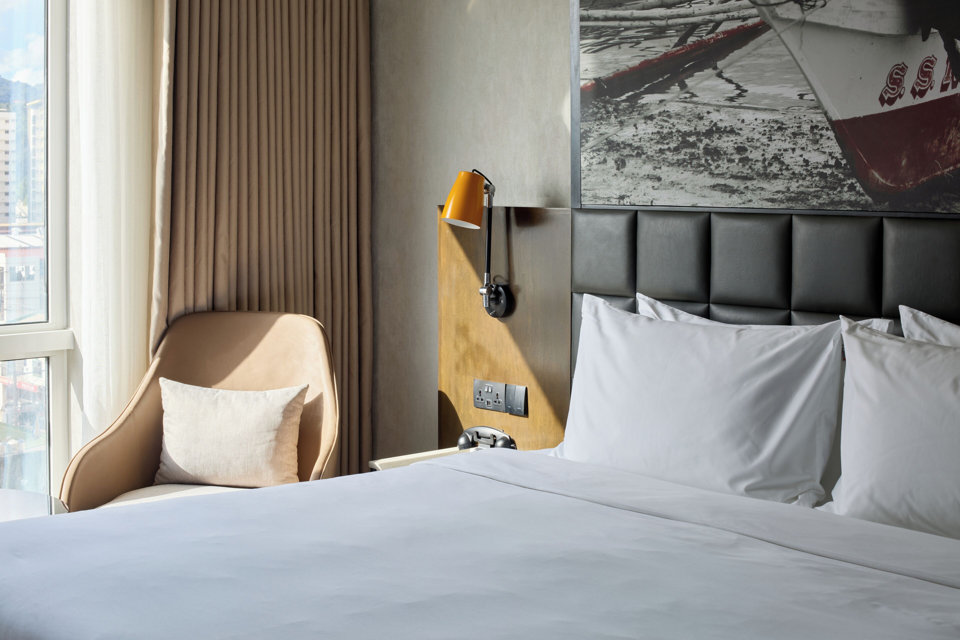 Habitación de hotel con cama, una silla, una lámpara de pared y un ventanal grande.