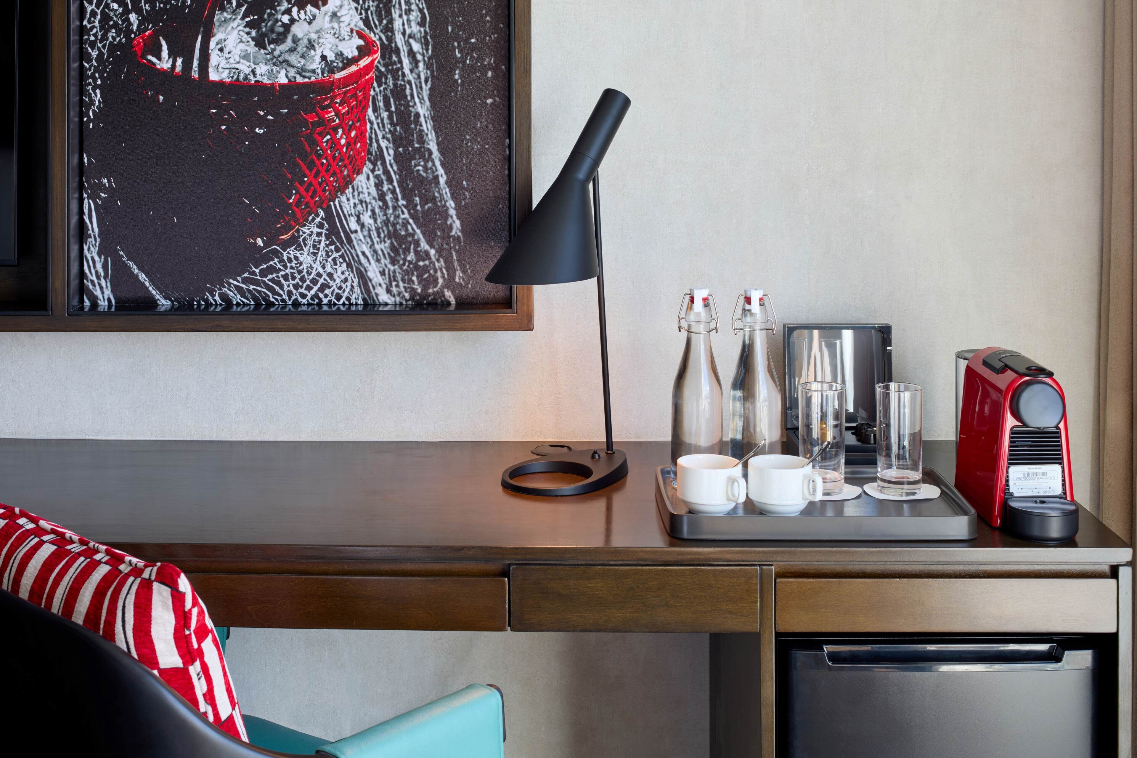 A hotel room with a wooden desk, a framed artwork, a black desk lamp, a coffee maker, and a tray with cups and glasses.