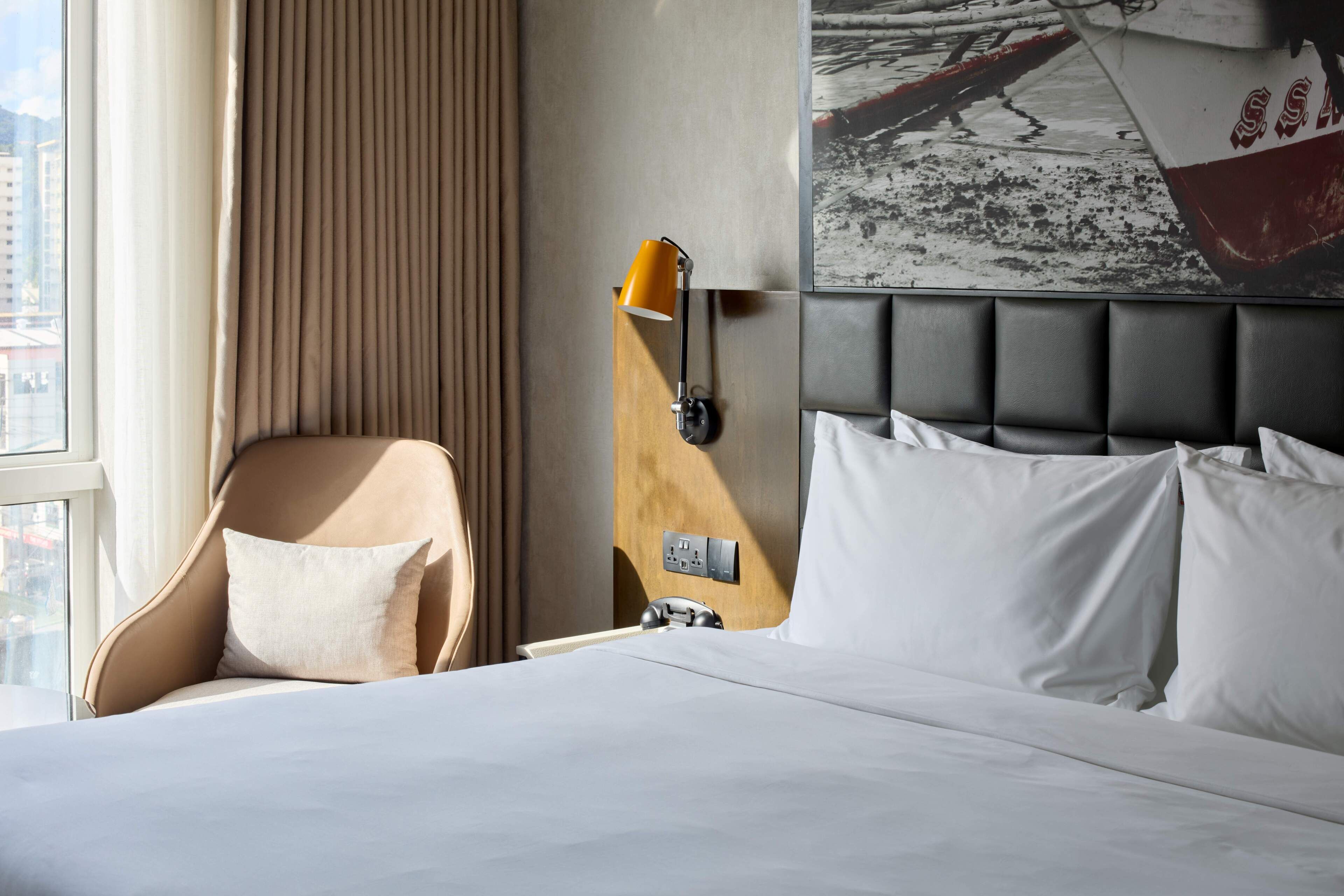 A hotel room with a bed, a chair, a wall-mounted lamp, and a large window.